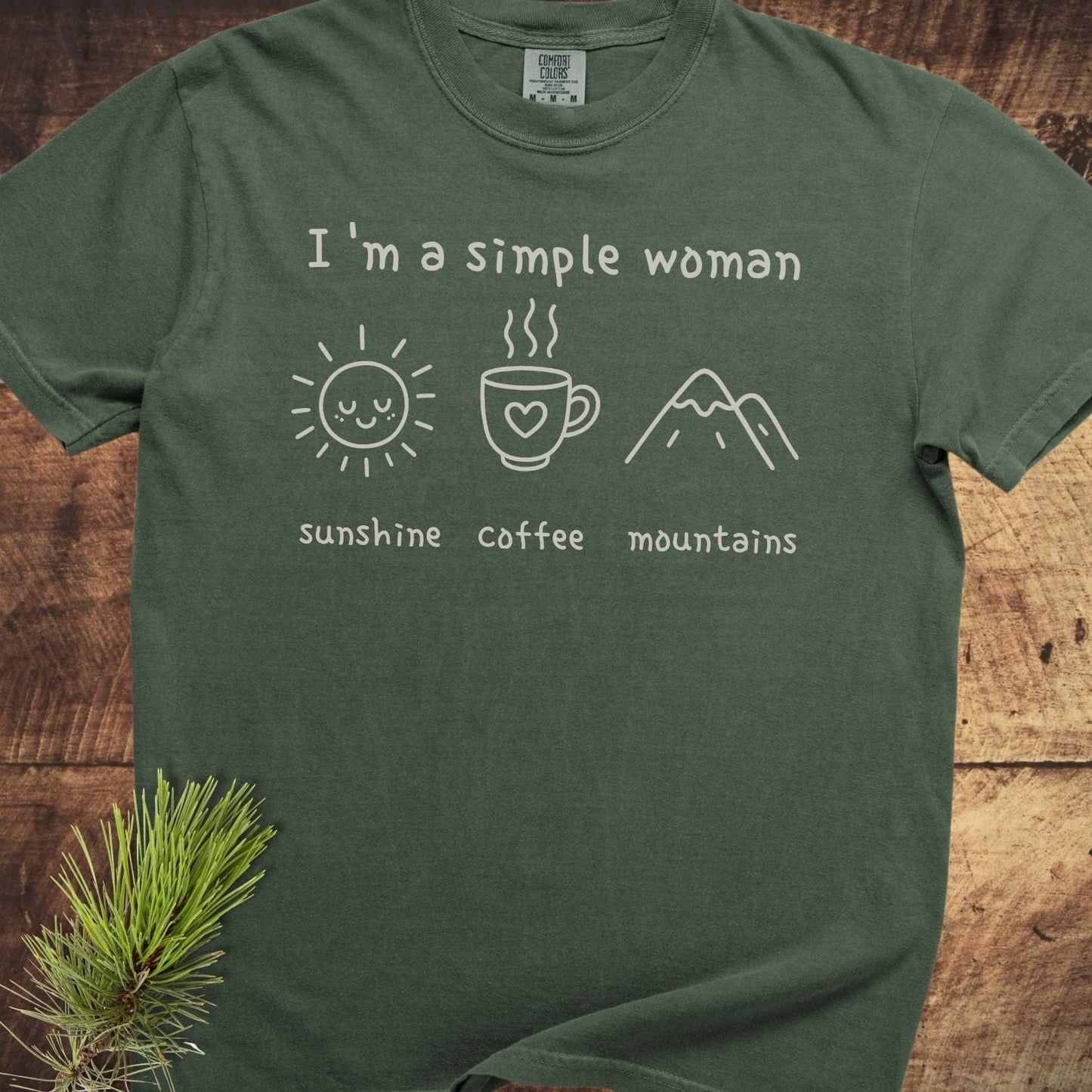 I'm A Simple Woman Comfort Colors 1717 Tee: forest-green, printed with sun, coffee, mountains icons. Soft-washed, relaxed fit, durable stitching, cotton fabric laid on wooden surface.