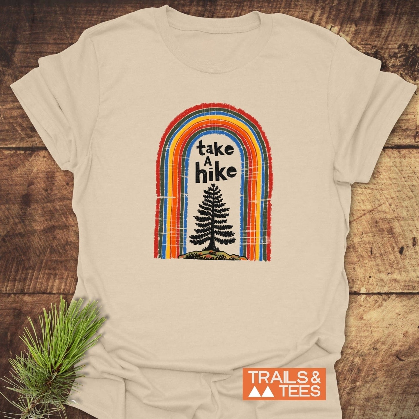 Take A Hike Rainbow T-Shirt on wood, featuring a retro arch and pine tree graphic with take a hike text. Includes Trails & Tees logo.