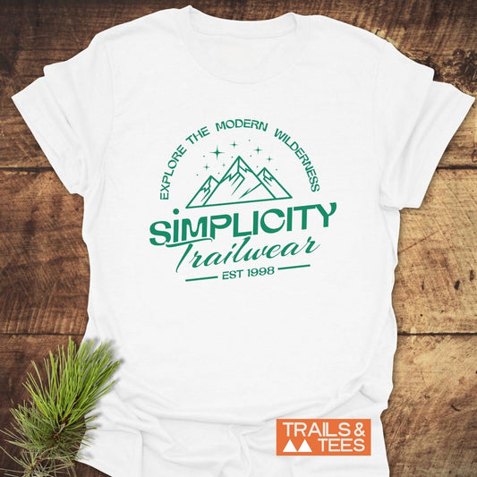 Simplicity Trailwear T-Shirt featuring a forest-green outdoor logo with text Explore the Modern Wilderness and EST 1998 on a wooden surface.