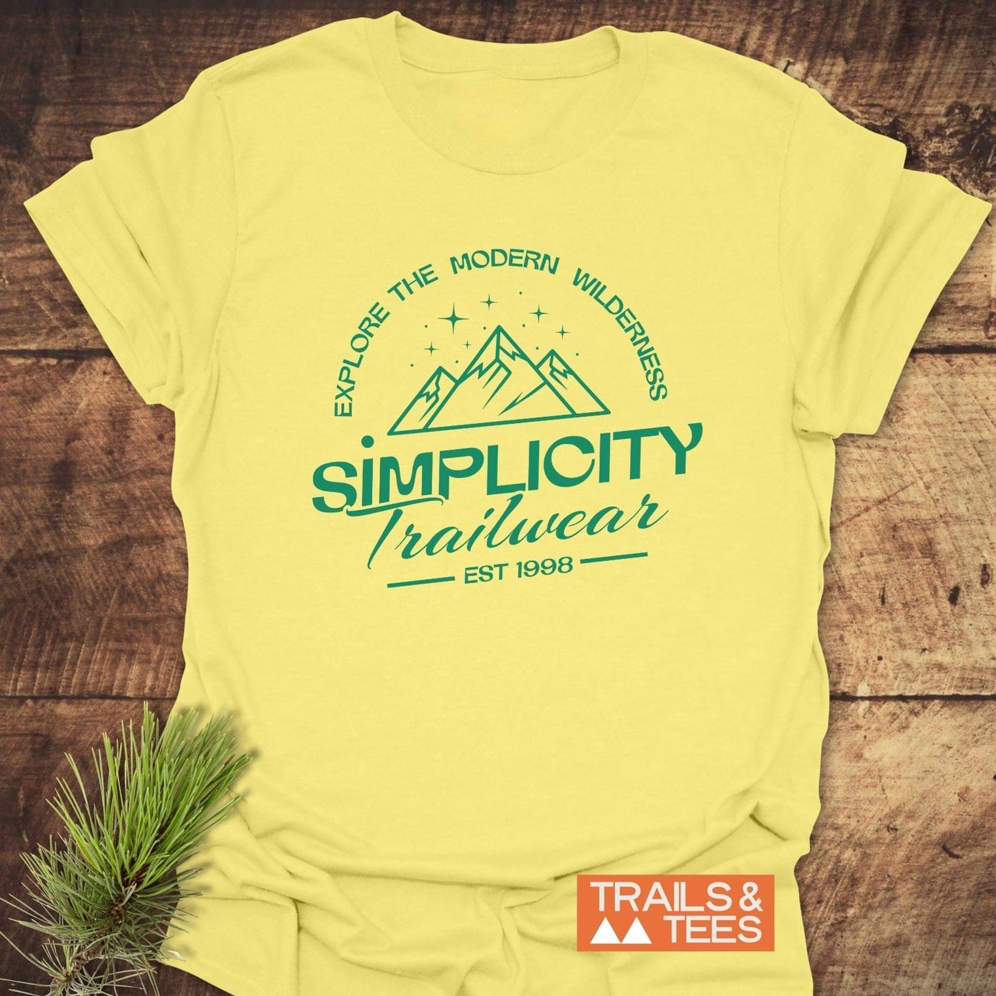Simplicity Trailwear T-Shirt featuring a mountain print with “EXPLORE THE MODERN WILDERNESS” text, laid on wood surface with pine sprig and “TRAILS & TEES” logo.