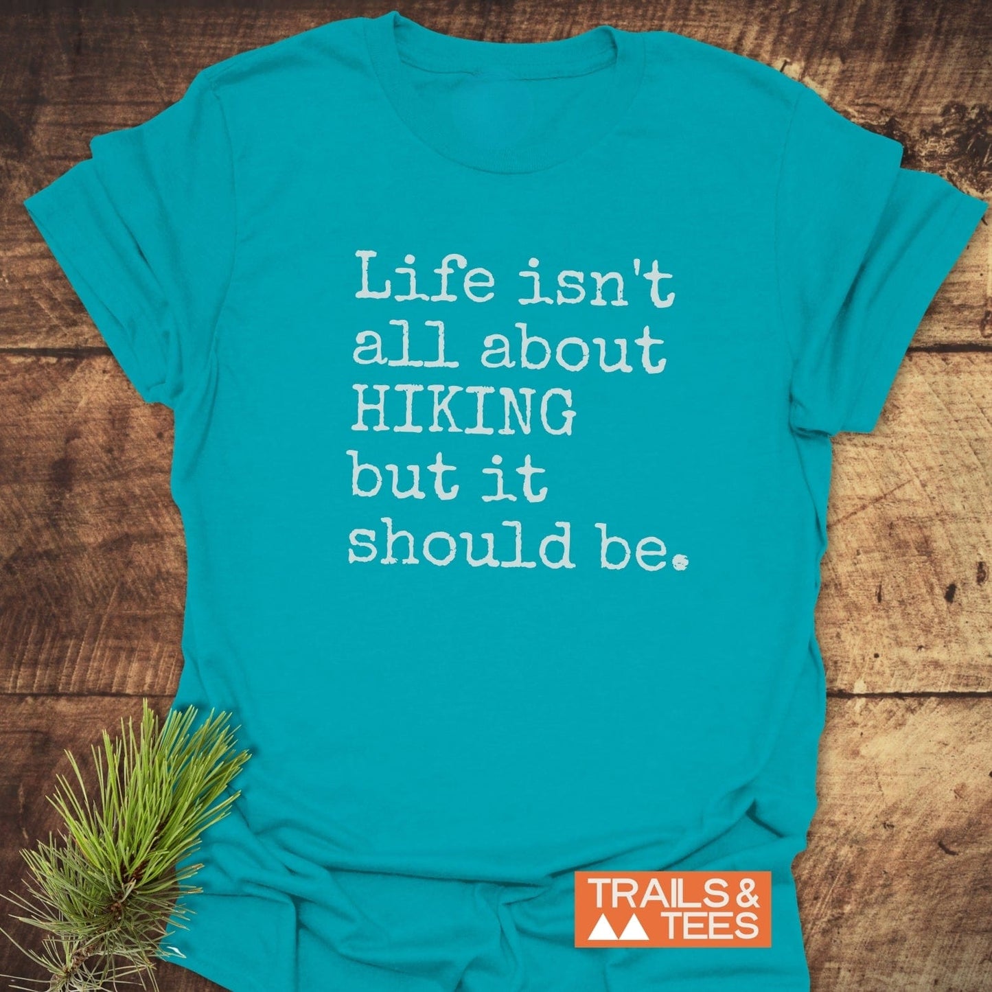 Teal Life Isn't All About Hiking T-Shirt with white typewriter-style text, laid on wooden surface, features Trails & Tees logo and pine sprig for style.