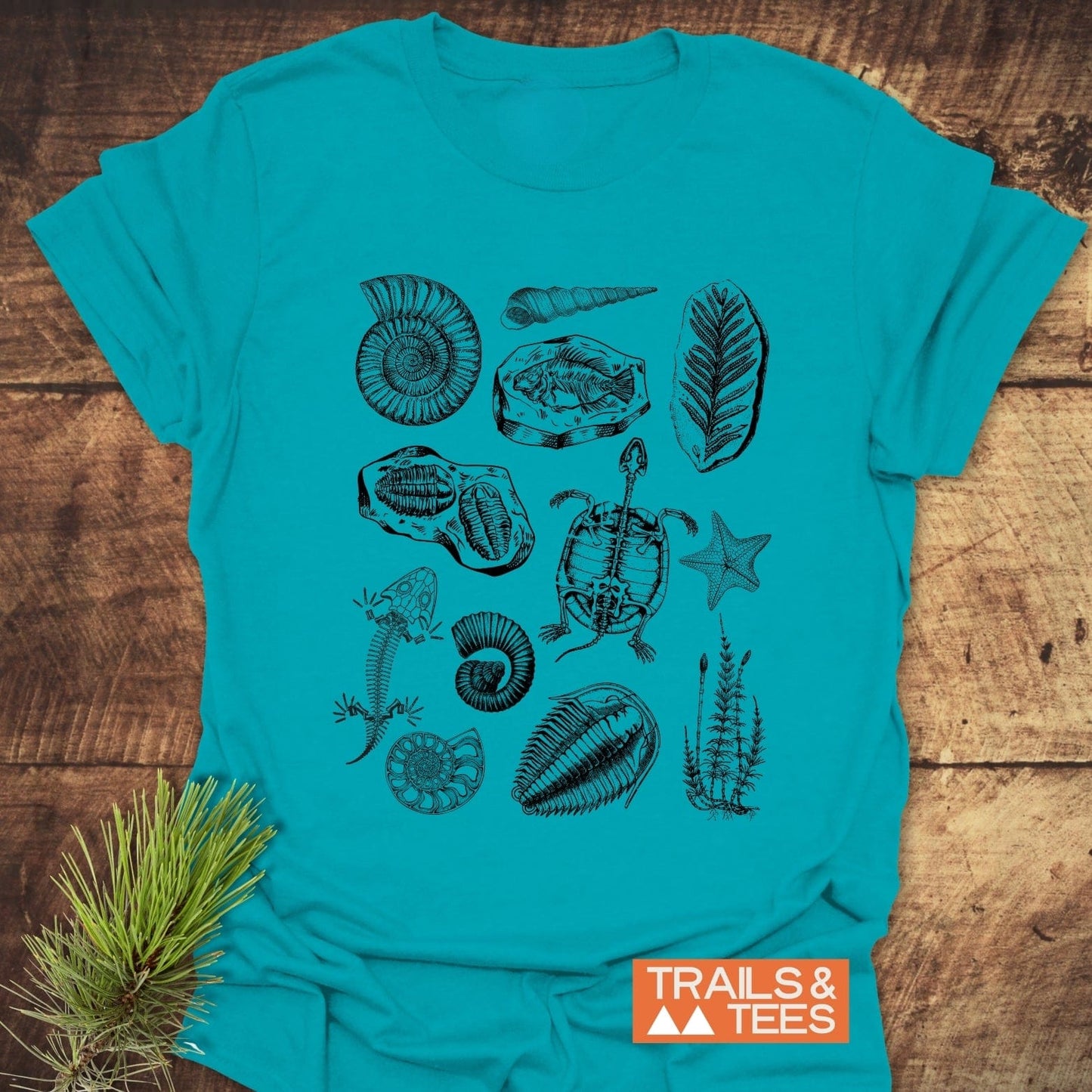 Fossile Vintage Tattoo T-Shirt featuring black fossil and marine life sketches, including ammonites and trilobites, laid on wood with TRAILS & TEES logo.