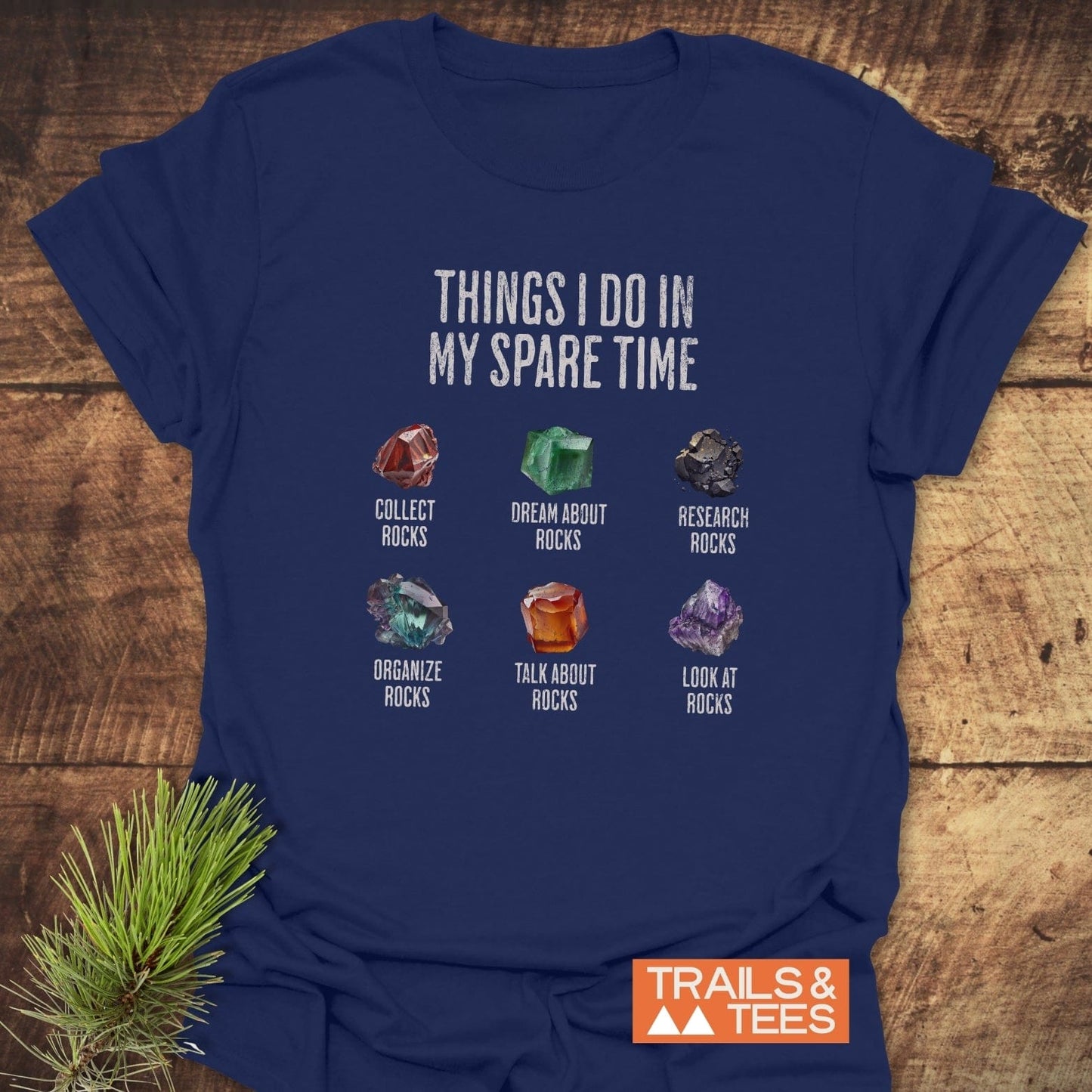 Sparetime Rock Lover T-Shirt featuring six rock illustrations with captions about rock collecting. Displayed with TRAILS & TEES logo on a wooden background.