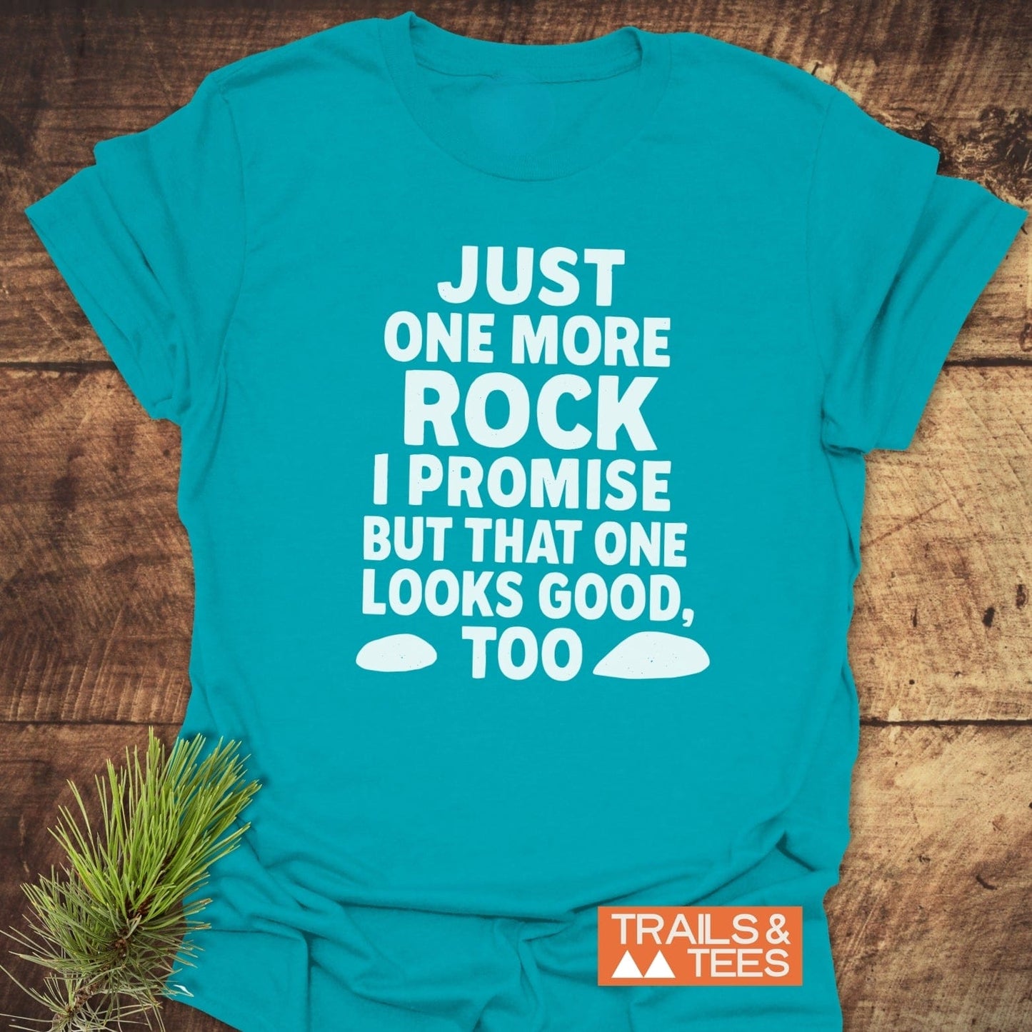 Just One More Rock Funny T-Shirt featuring a bold slogan with rock icons and a Trails & Tees logo, laid flat with a pine sprig beside it.