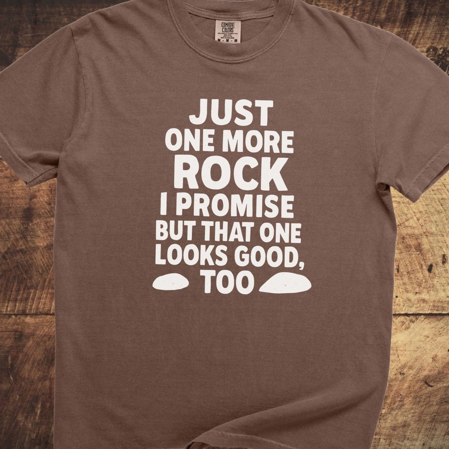 Just One More Rock Funny Comfort Colors 1717 Tee with humorous white text and rock shapes, laid on a wooden surface, showcasing its relaxed fit and durable stitching.