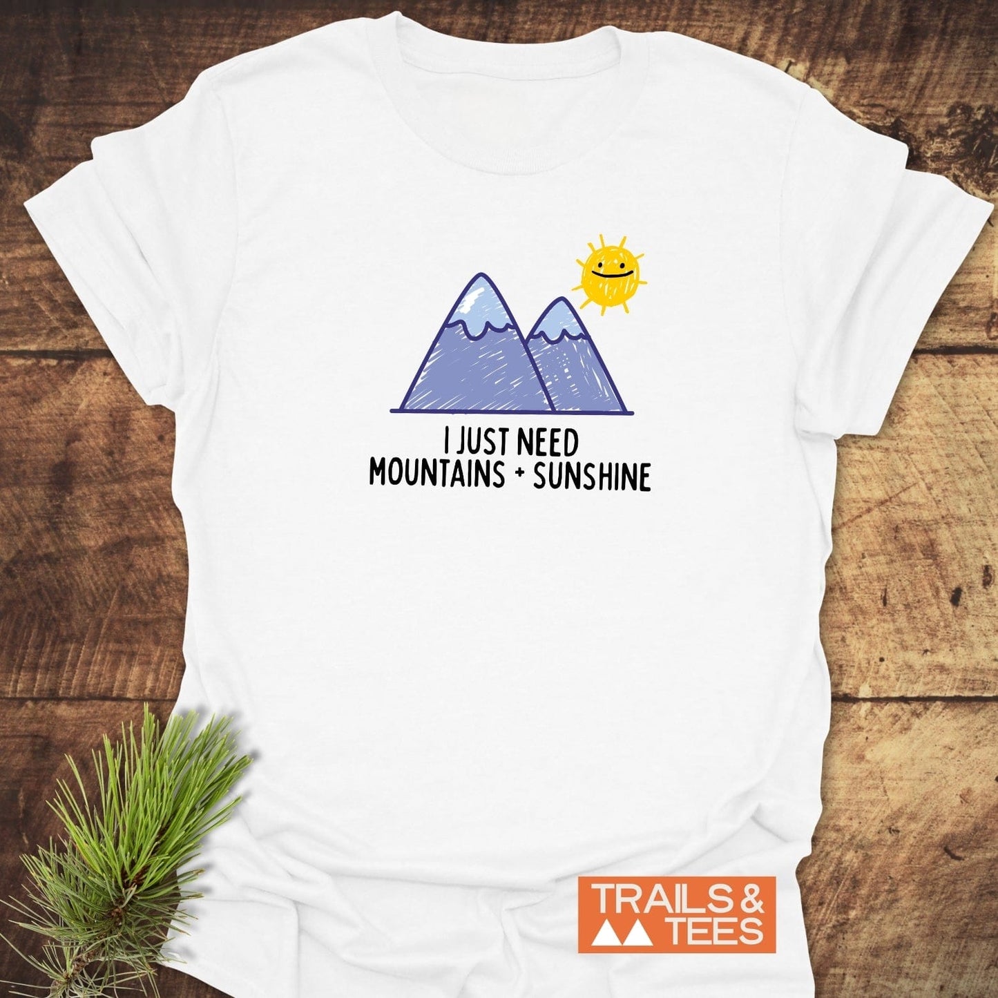 Mountains+Sunshine T-Shirt featuring a hand-drawn graphic of purple mountains and a smiling sun, with the slogan “I JUST NEED MOUNTAINS + SUNSHINE.”