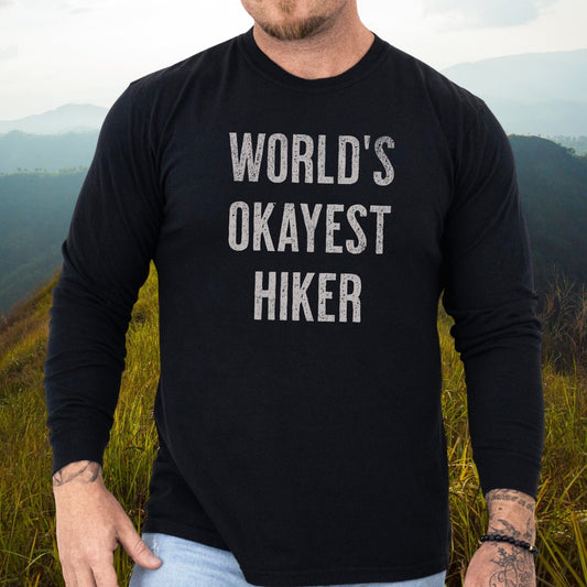 Person in hilly landscape wearing World's Okayest Hiker black long-sleeve tee, showcasing tattoos and bead bracelet, paired with light jeans.