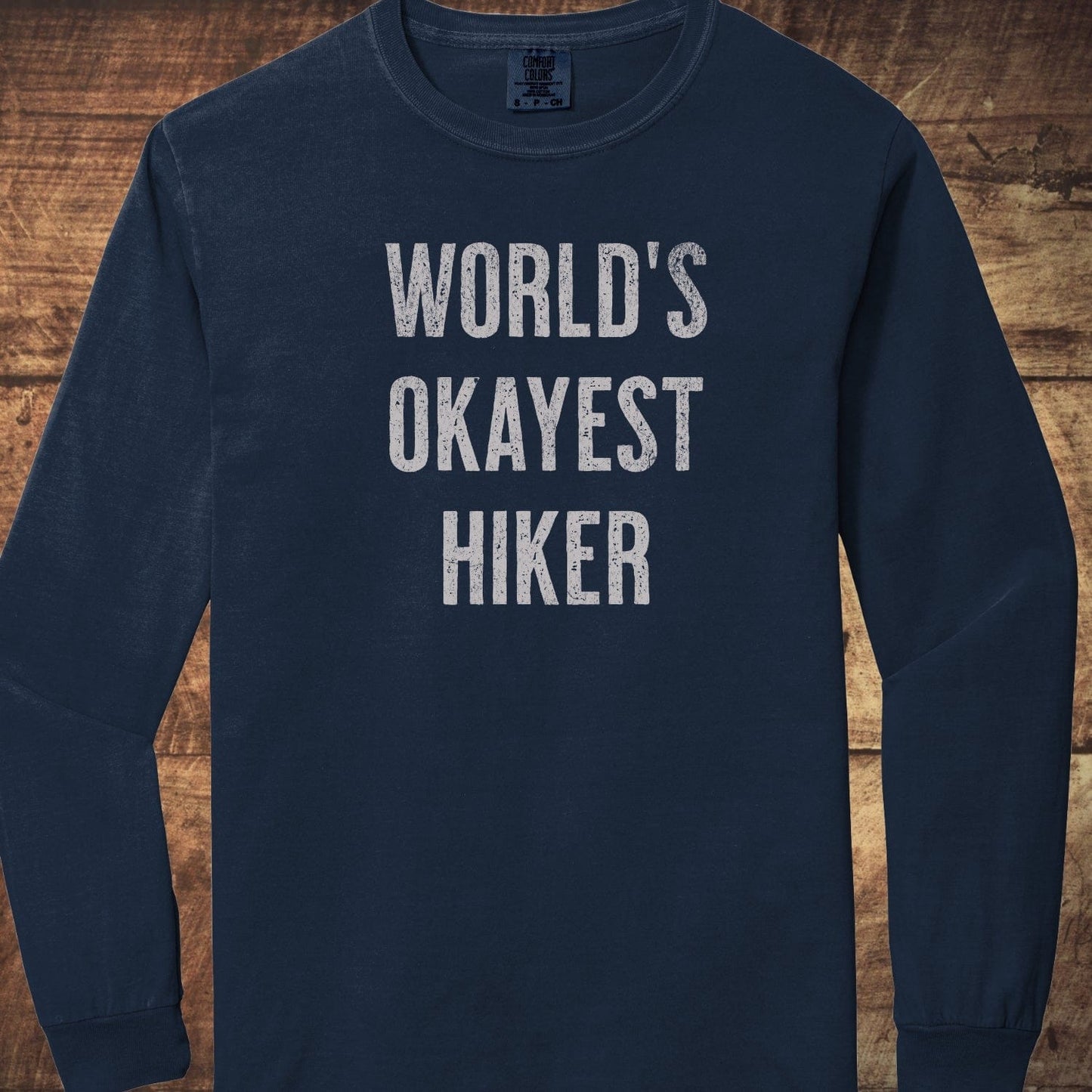 World's Okayest Hiker Comfort Colors Long-Sleeve Tee, navy-blue, with white text, laid on wooden background; 100% ring-spun cotton, relaxed fit, garment-dyed fabric.