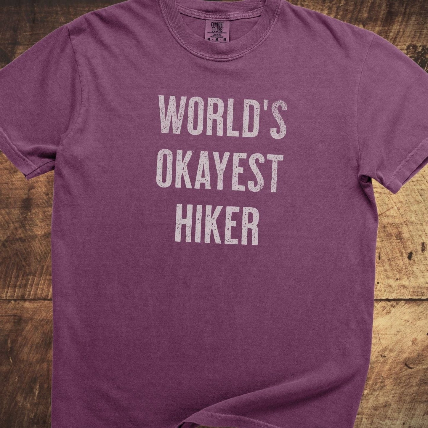 World's Okayest Hiker Comfort Colors 1717 Tee in purple heathered fabric, featuring distressed white text, soft-washed cotton, relaxed fit, laid on wood surface.