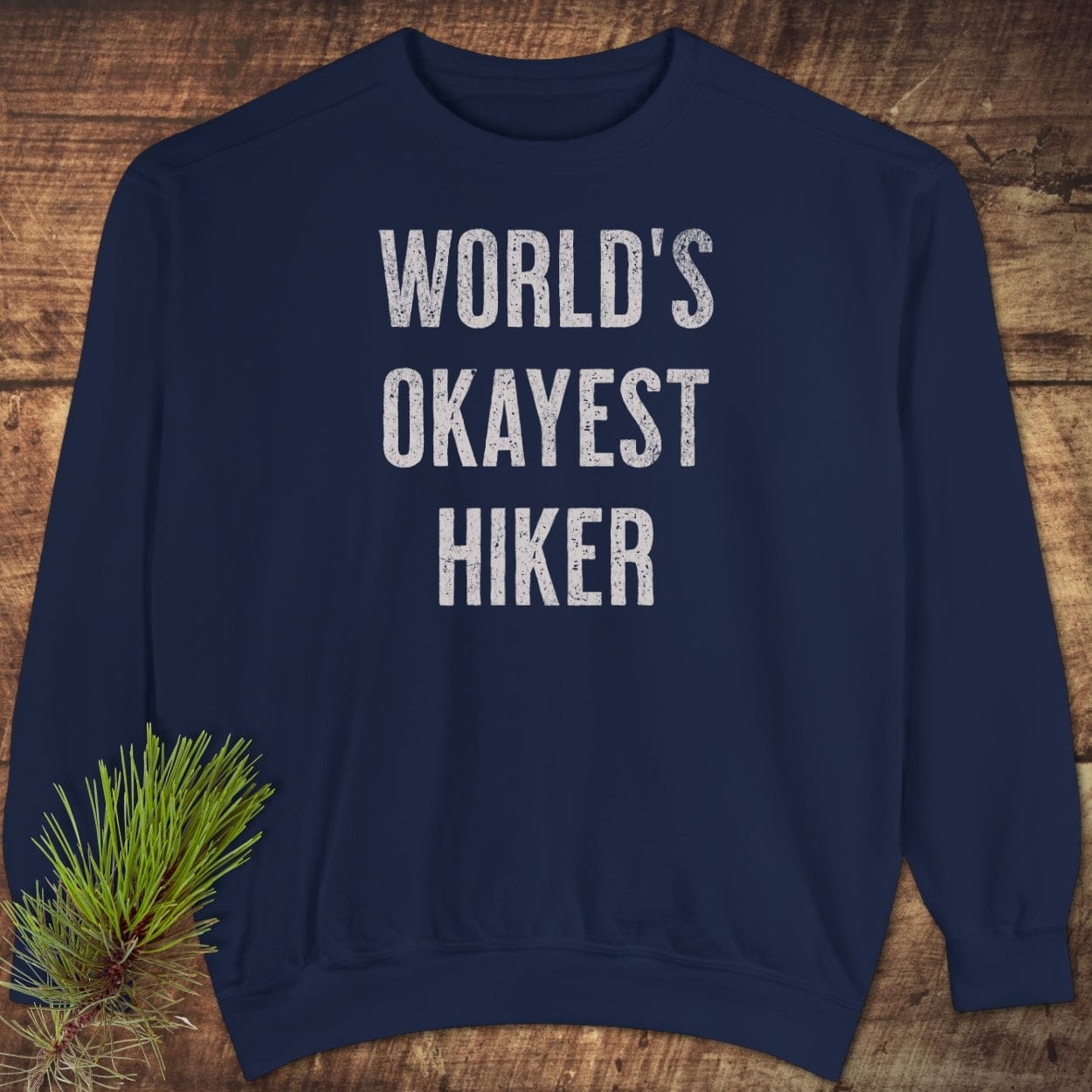 Navy-blue World's Okayest Hiker sweatshirt on wood, featuring distressed white text, relaxed fit, and rustic pine accent for an outdoorsy touch.