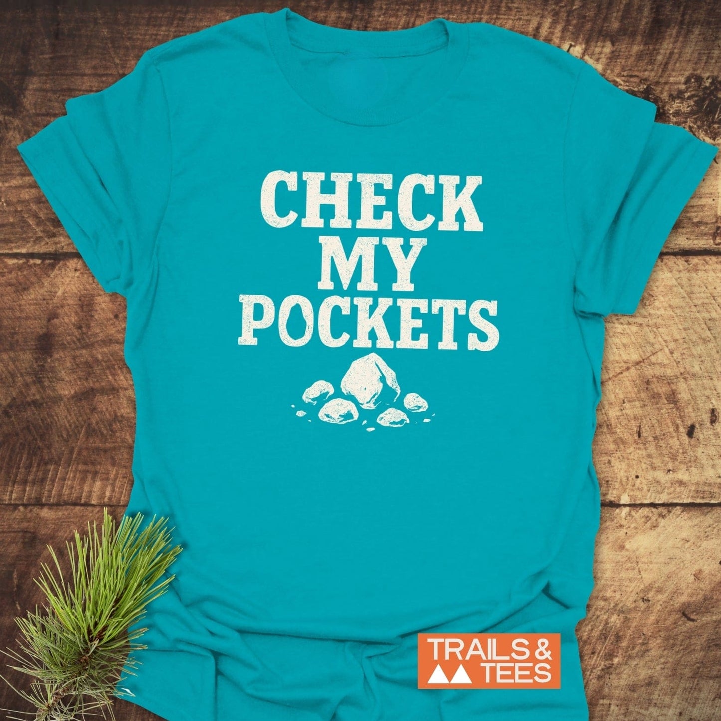 Check My Pockets Rock Lover T-Shirt with white print, rocks graphic, and Trails & Tees label, on wooden surface with pine sprig.