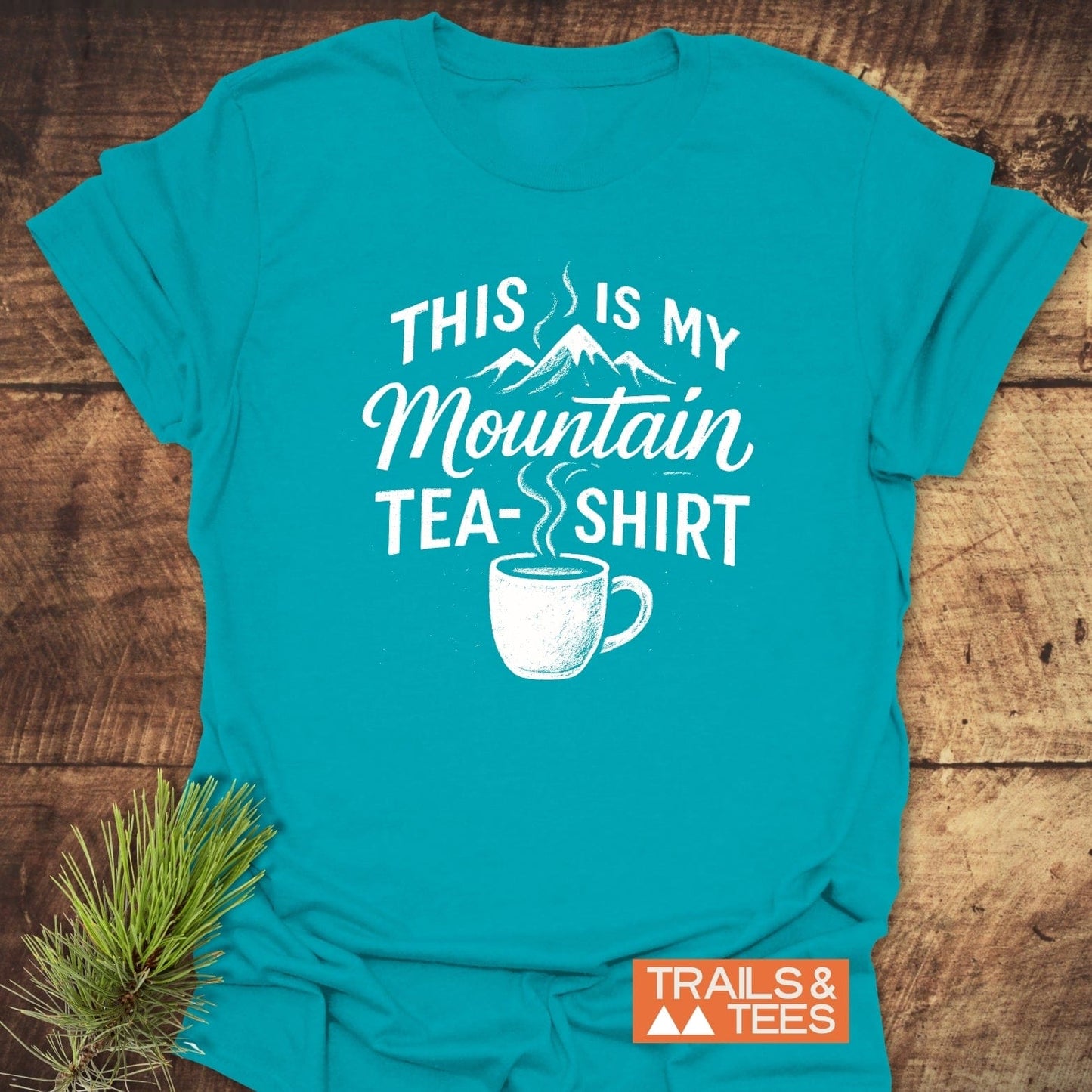 Mountain Tea T-Shirt featuring a mountain graphic and slogan, laid on wood with pine sprig; crafted from soft, pre-shrunk cotton for outdoor enthusiasts.