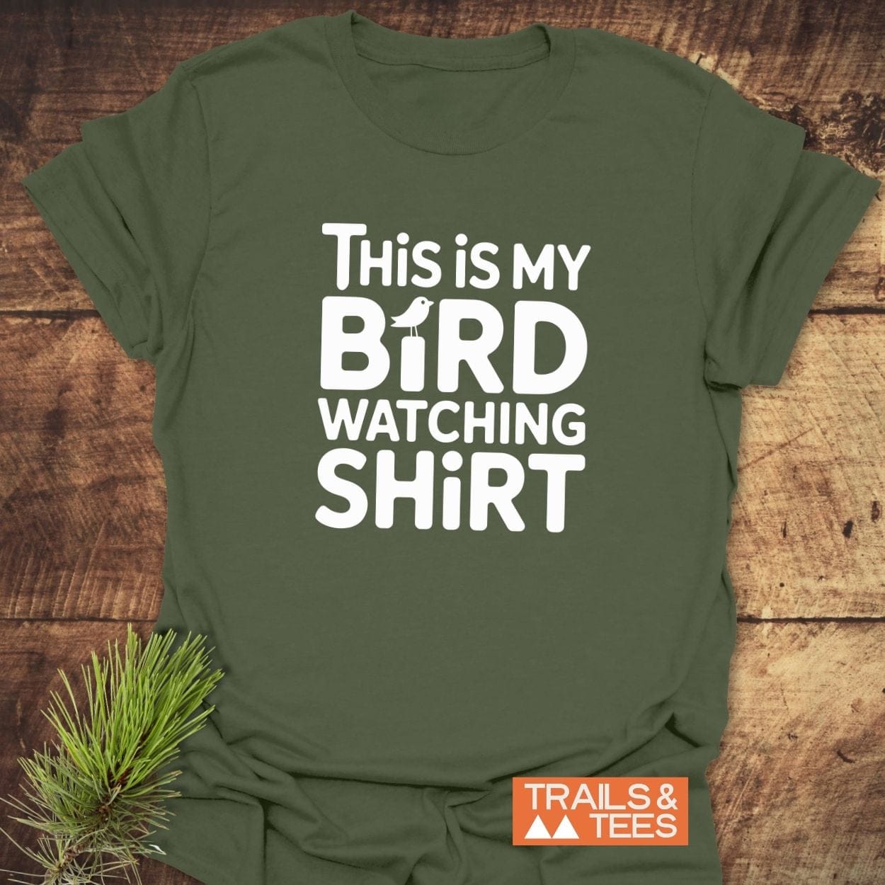 Olive-green Bird Watching T-Shirt with bold white text and bird silhouette, laid on wood, Trails & Tees logo visible, pine sprig tucked under hem.