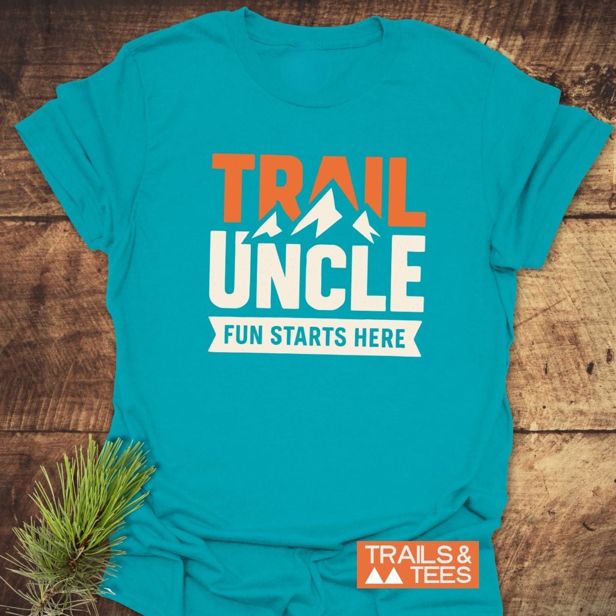 Trail Uncle Funny T-Shirt with a graphic featuring mountain peaks and bold text. Laid on wood with a pine sprig beside it.