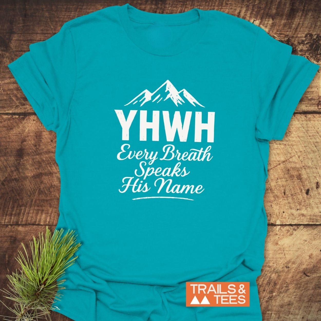 YHWH Christian Hiking T-Shirt with mountain silhouette and spiritual text, crafted from soft cotton for comfort and durability, featuring a small “TRAILS & TEES” logo.