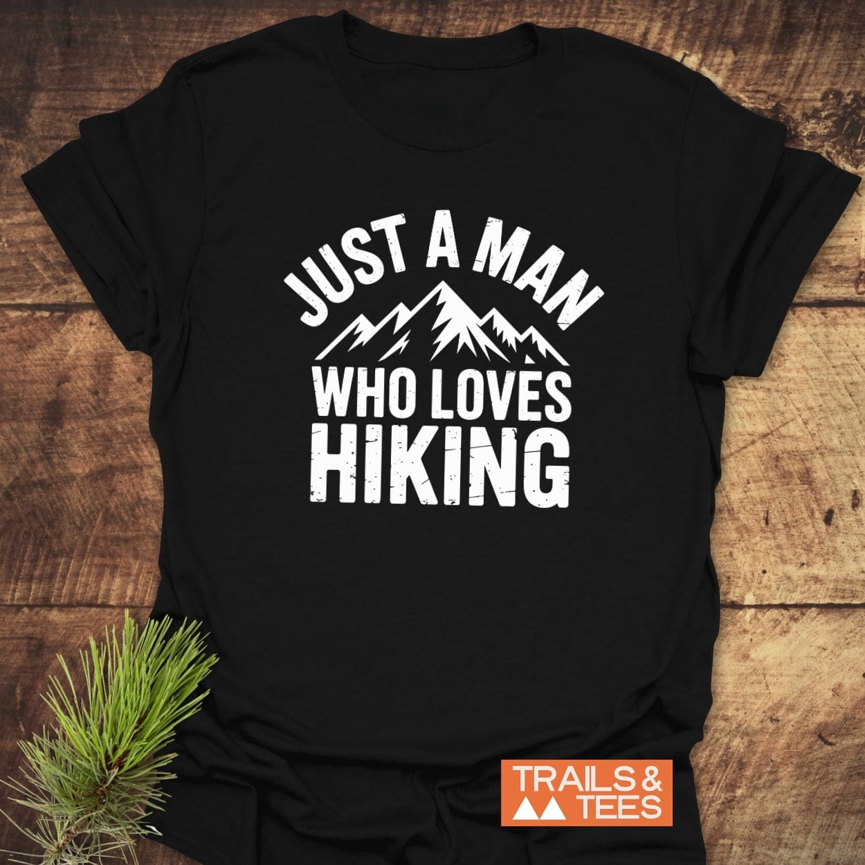 Just A Man... Hiking T-Shirt with white slogan and mountain graphic, featuring Trails & Tees logo, laid on wood with pine sprig for a timeless outdoor look.