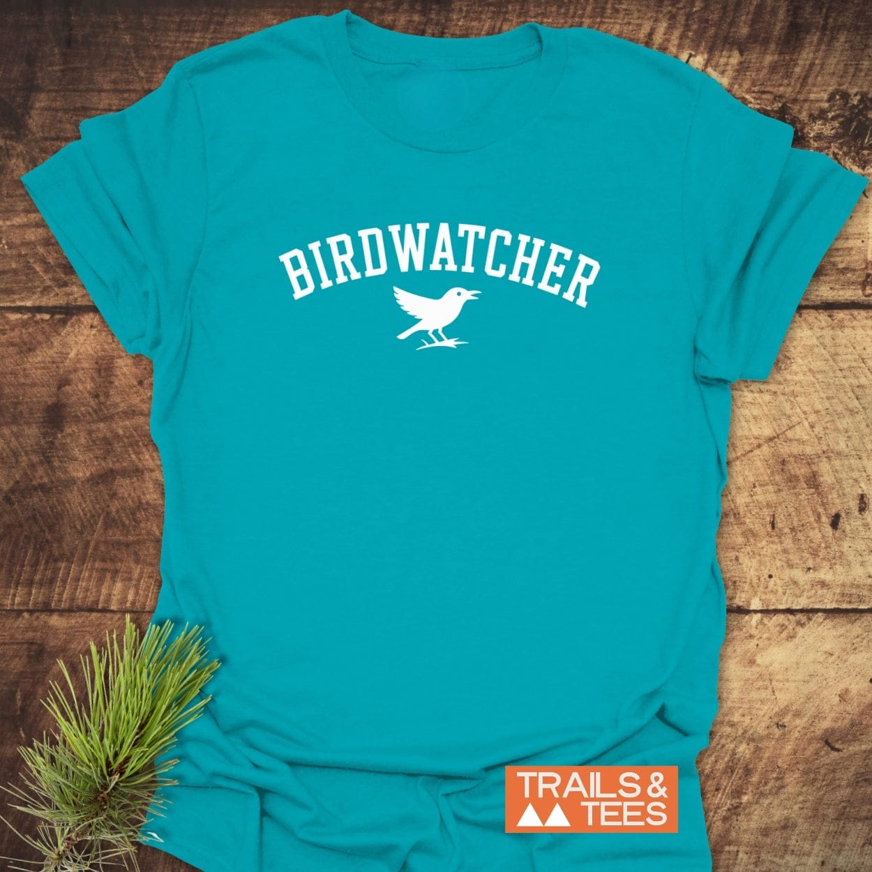 Birdwatcher Varsity T-Shirt on wood, featuring bold lettering and bird silhouette, highlighting its outdoor theme and brand logo, Trails & Tees.