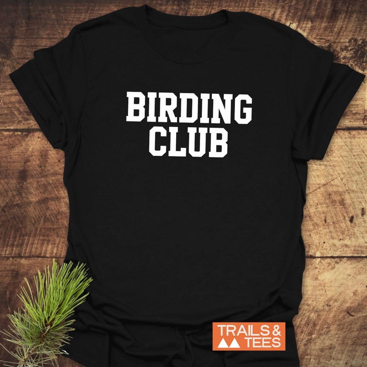 Birding Club T-Shirt on wood surface, featuring bold white text. Soft, lightweight cotton for outdoor adventures. Trails & Tees logo visible in corner.