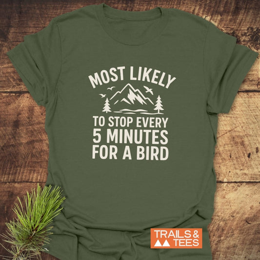 Most Likely Bird Watcher T-Shirt features a mountain scene print, slogan, and orange label, showcasing its outdoor-inspired design on a wooden surface.