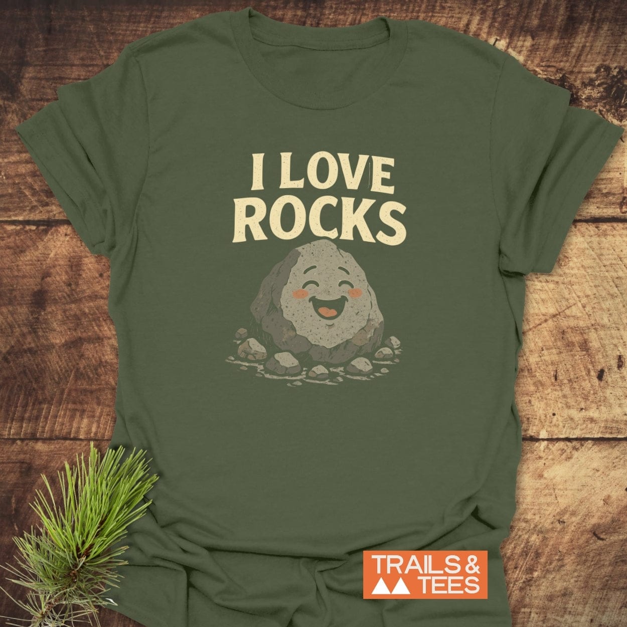 I Love Rocks T-Shirt on wood surface; features a cartoon boulder and stones. Made of soft, lightweight cotton with durable print, designed for outdoor enthusiasts.