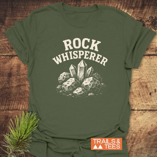 Rock Whisperer T-Shirt featuring a crystal graphic and ROCK WHISPERER text, displayed on a wooden surface with Trails & Tees logo and pine sprig accent.