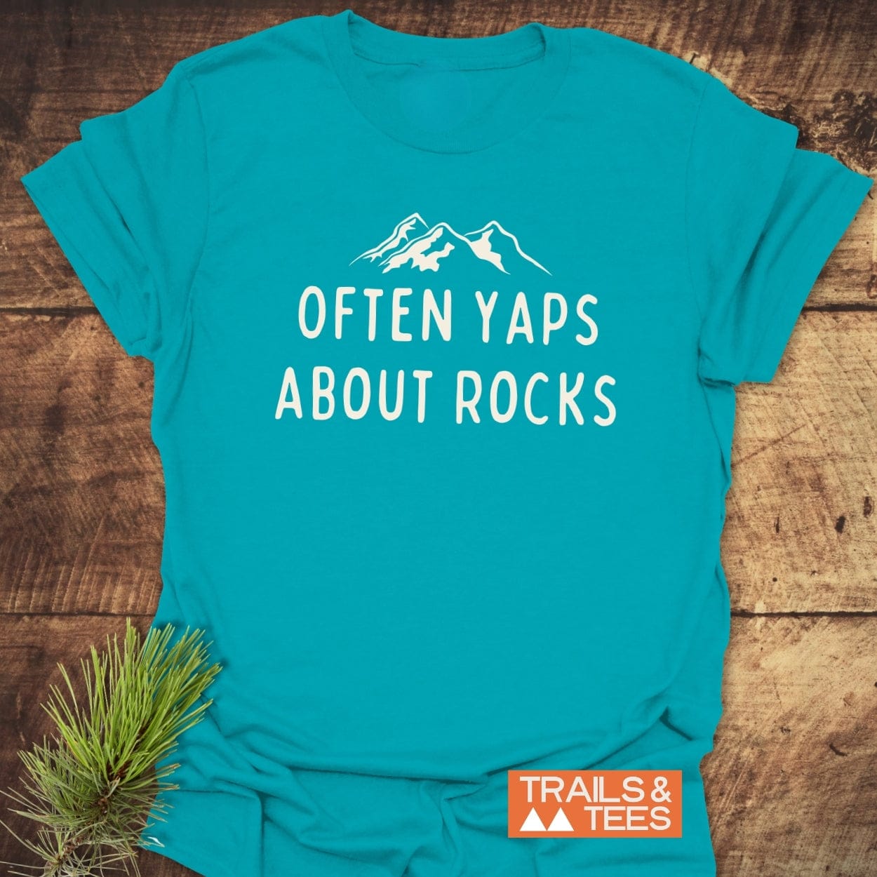 Often Yaps About Rocks T-Shirt with mountain silhouette; teal cotton, soft, durable, pre-shrunk, features Trails & Tees logo, ideal for outdoor enthusiasts.