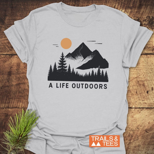 A flat-lay of the A Life Outdoors T-Shirt featuring mountain and sun graphic, pine tree illustration, and slogan, emphasizing adventure and outdoor lifestyle.