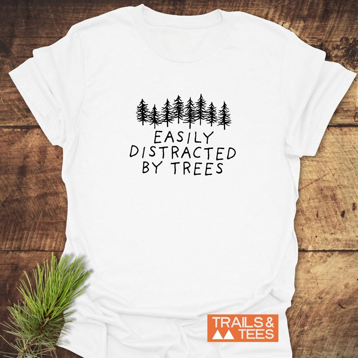 Easily Distracted By Trees T-Shirt with pine tree graphic, made from soft ring-spun cotton, laid flat with a pine sprig and Trails & Tees logo nearby.