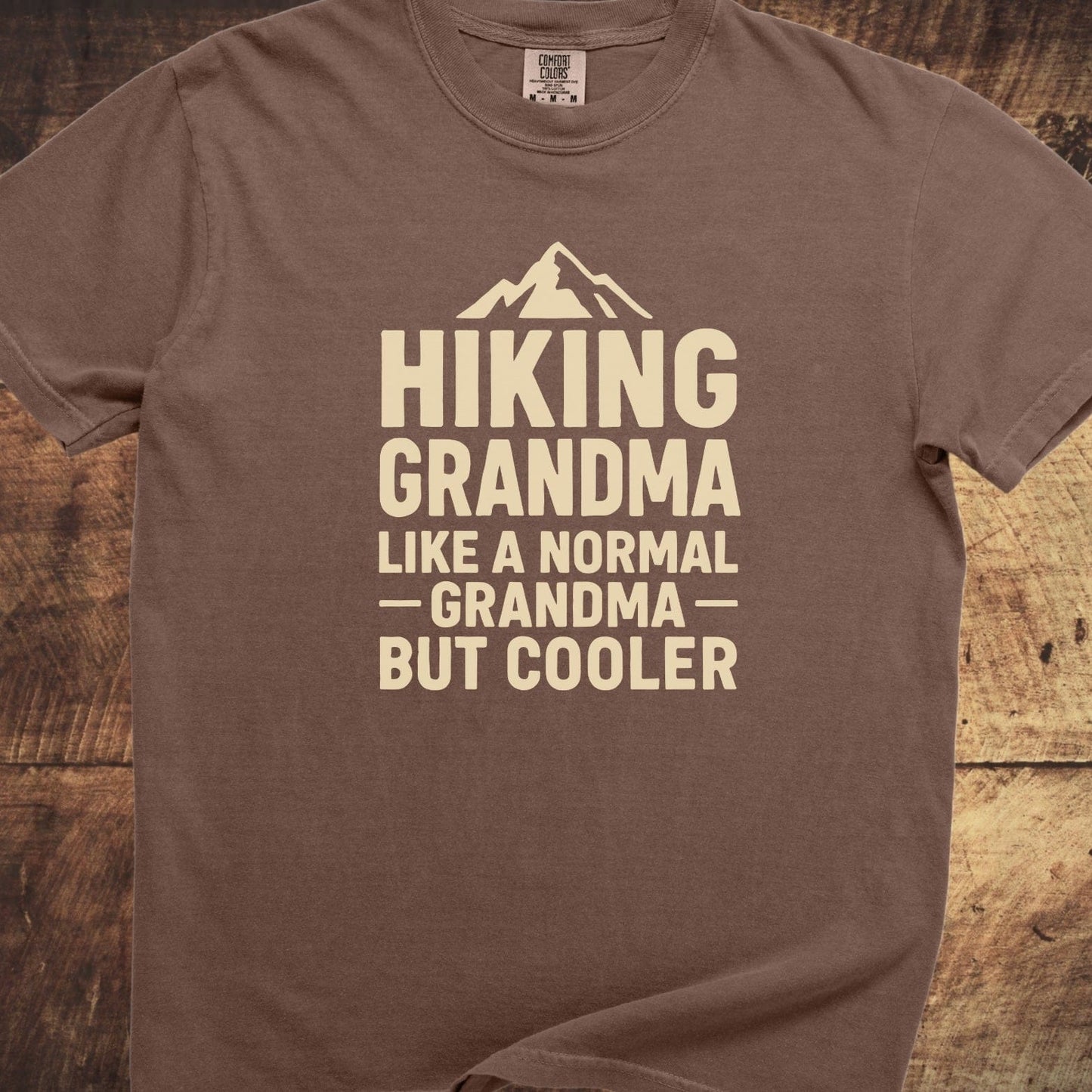 Hiking Grandma Comfort Colors 1717 Tee features a mountain graphic and text on a brown, short-sleeve T-shirt, laid on a wooden surface.