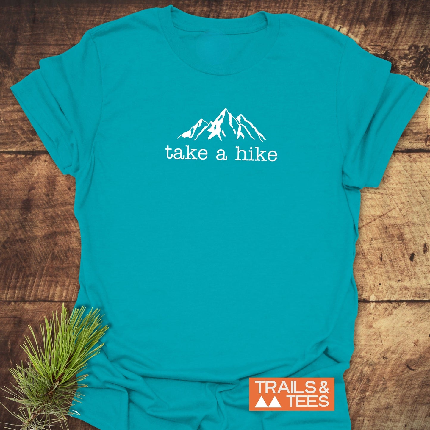 Take A Hike T-Shirt featuring a white mountain line-drawing and take a hike text, displayed on a wooden surface with a small orange TRAILS & TEES badge and pine needles nearby.