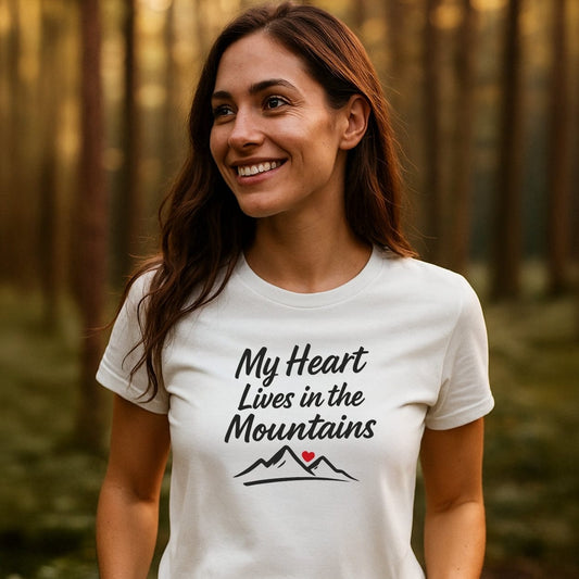 Young woman wearing the My Heart Lives In The Mountains T-Shirt in a forest at sunset, showcasing its stylized script and mountain design, capturing the essence of outdoor adventure and comfort.