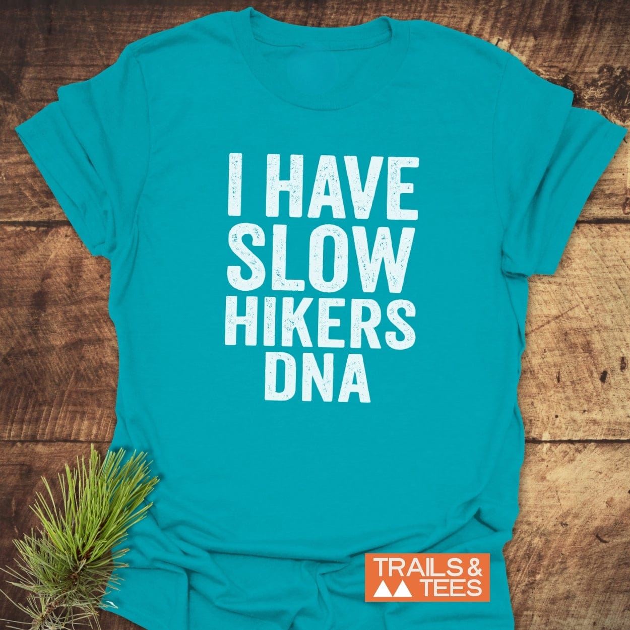 Slow Hikers DNA T-Shirt with white block letters on turquoise-blue fabric, displayed flat on wood with Trails & Tees logo and pine branch accent, highlighting its outdoor-inspired design and comfort.