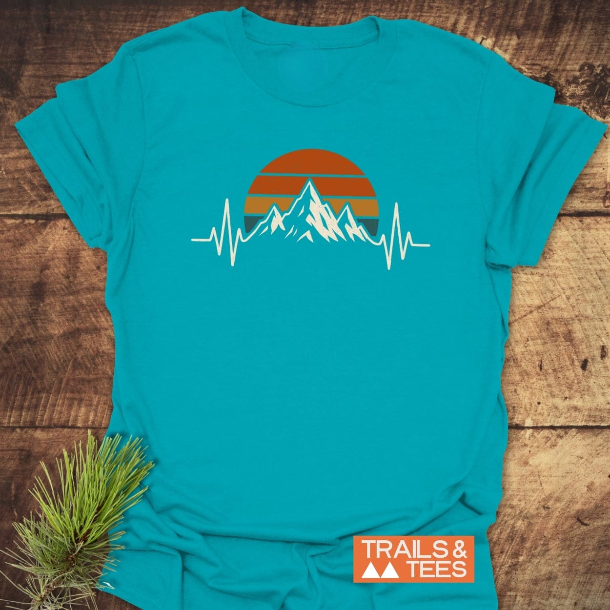 Mountain Heartbeat T-Shirt with jagged mountain peaks and a stylized heartbeat line design on chest, laid flat on wood surface. Displayed with Trails & Tees logo and pine sprig alongside.