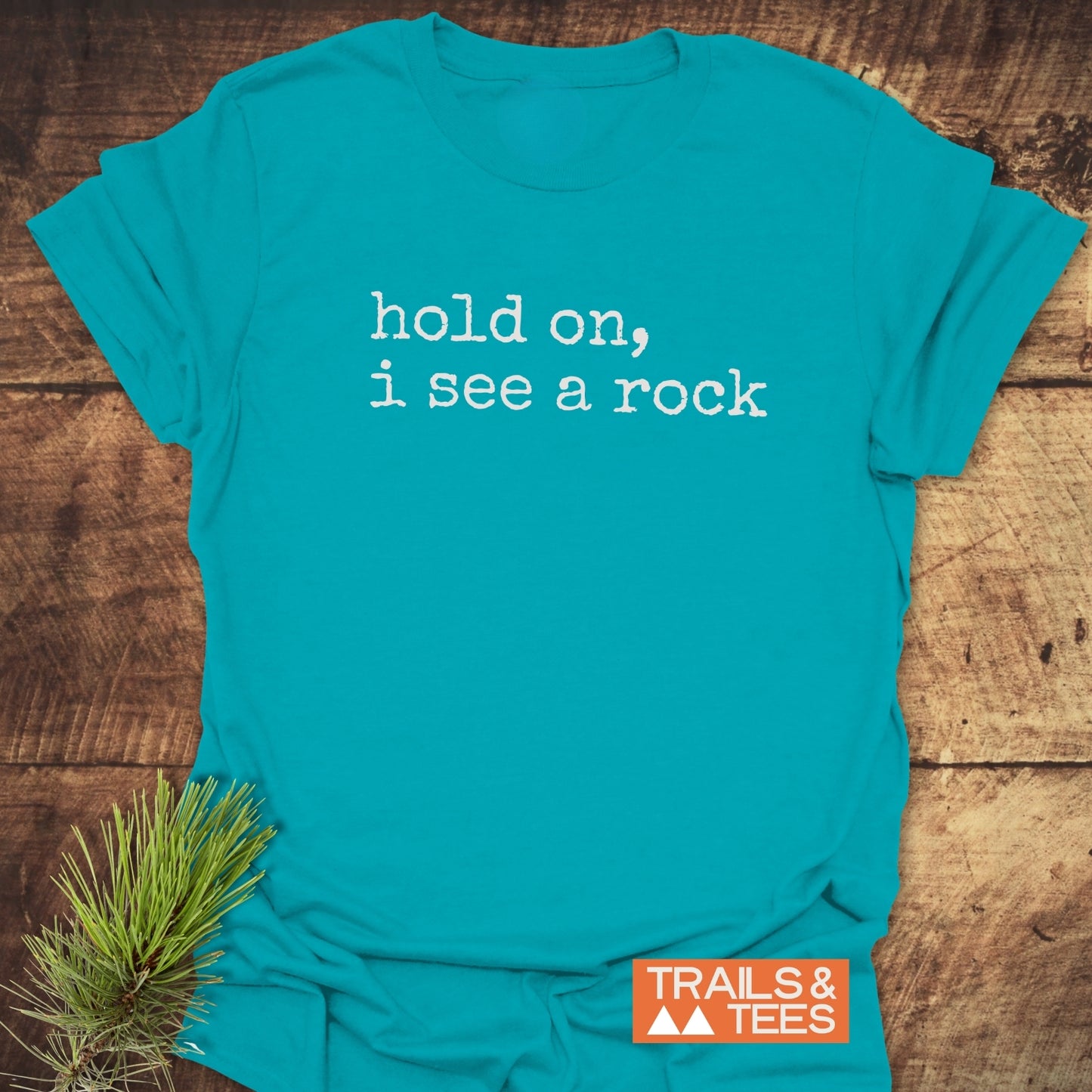 Hold On I See A Rock T-Shirt flat-lay on rustic wood, featuring distressed white typewriter-style text. Accented with a pine branch, highlighting its adventure-ready, soft ring-spun cotton design by TRAILS & TEES.