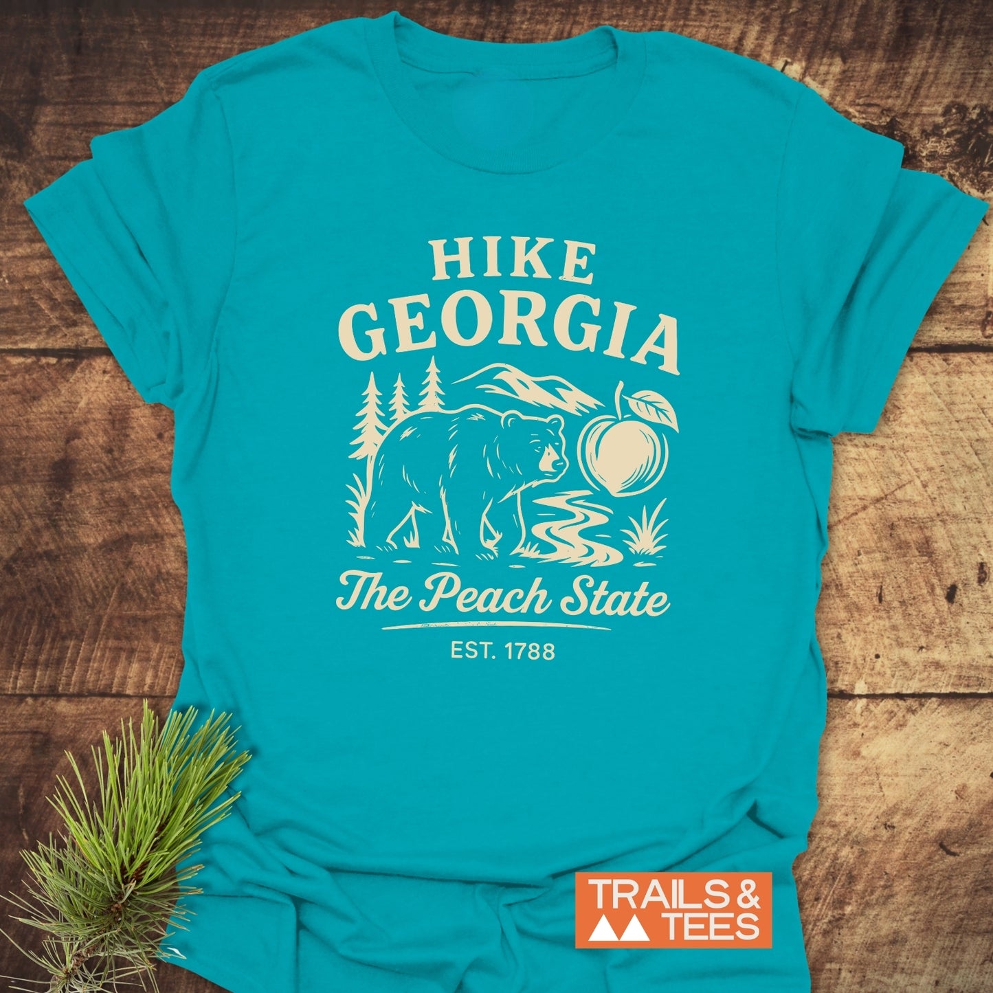 Hike Georgia T-Shirt featuring a bear by a stream, pine trees, and mountains with The Peach State and EST. 1788. Includes TRAILS & TEES logo, made for comfort and adventure.