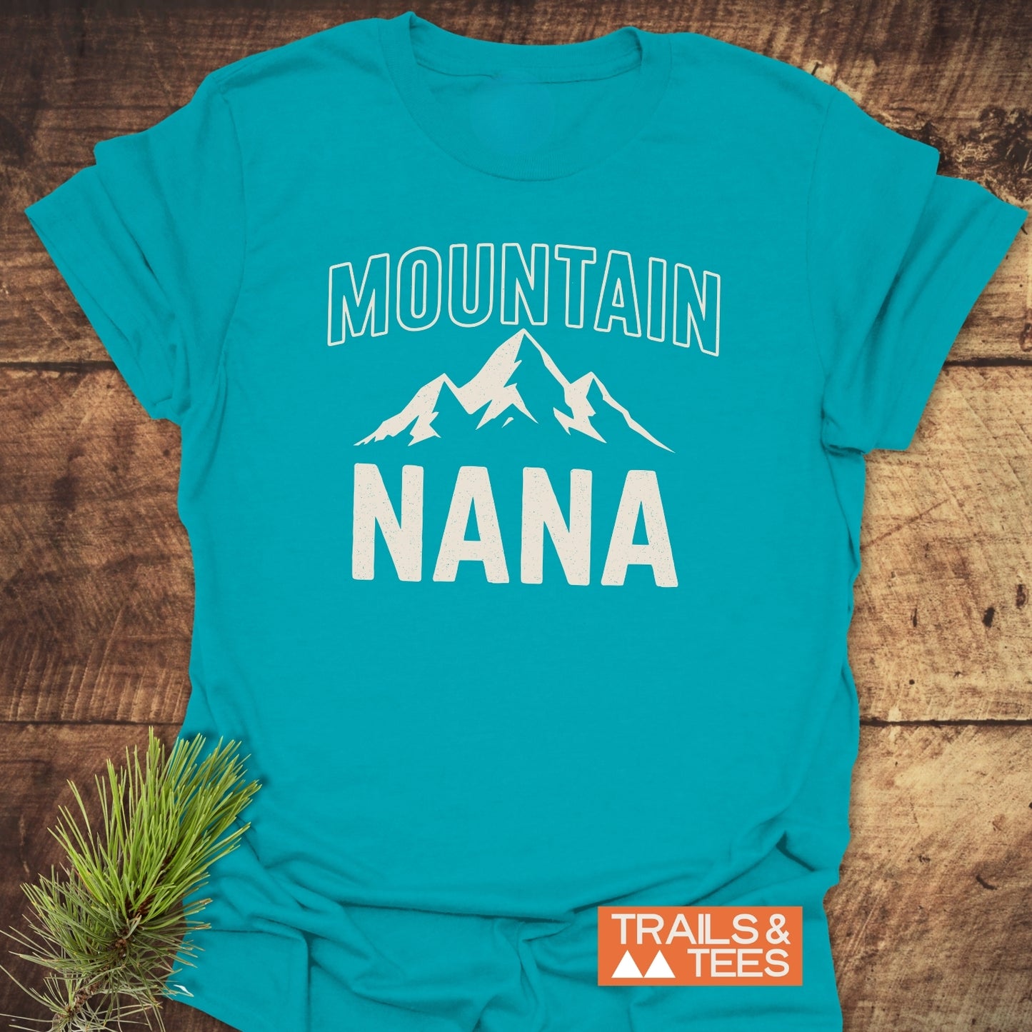 Mountain Nana T-Shirt featuring a white mountain graphic, “MOUNTAIN” above, “NANA” below, on a turquoise-blue shirt. Includes TRAILS & TEES logo and pine sprig accent.