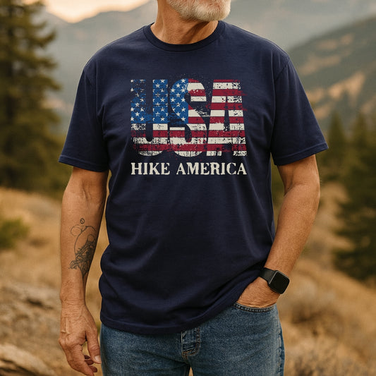 Man outdoors wearing Hike America T-Shirt with distressed American-flag motif spelling USA and HIKE AMERICA text, showcasing casual style and comfort, complemented by a smartwatch and tattoo.