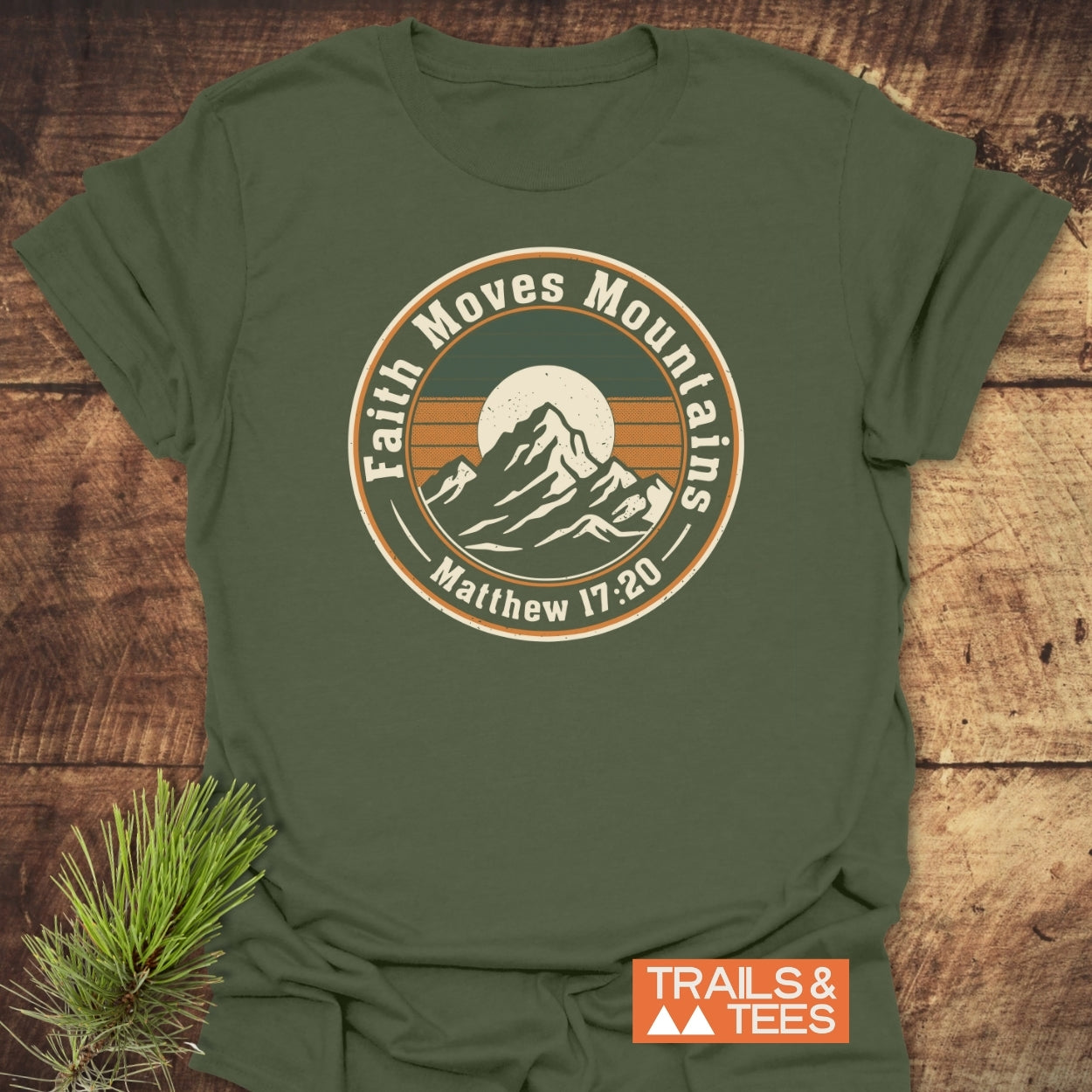 Faith Moves Mountains T-Shirt with a vintage mountain graphic and Bible verse, featuring a “TRAILS & TEES” logo; complemented by a pine sprig detail, highlighting outdoor spirit and adventure-ready comfort.