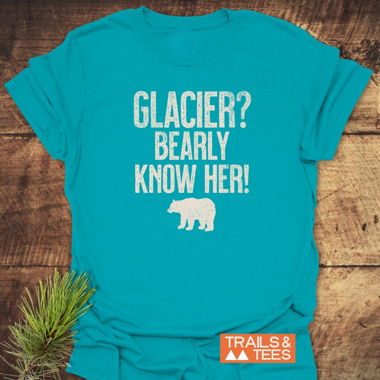 Glacier National Park Funny T-Shirt showcasing a white bear silhouette and humorous text, GLACIER? BEARLY KNOW HER! on a turquoise-blue backdrop, laid flat with Trails & Tees logo nearby.