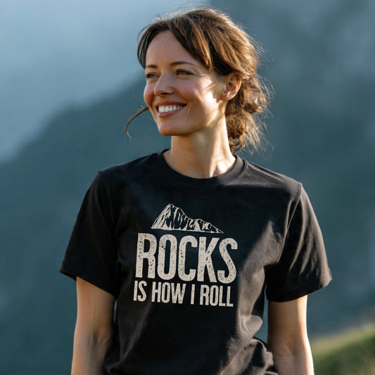 A woman outdoors wearing the Rocks Is How I Roll T-Shirt, showcasing a stylized mountain design and bold text, embodying adventure-ready comfort and durability for outdoor enthusiasts.
