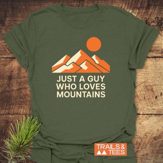 Mountain Love T-Shirt features a stylized mountain and sun print with “JUST A GUY WHO LOVES MOUNTAINS” text, laid flat on wood. Includes “TRAILS & TEES” logo.