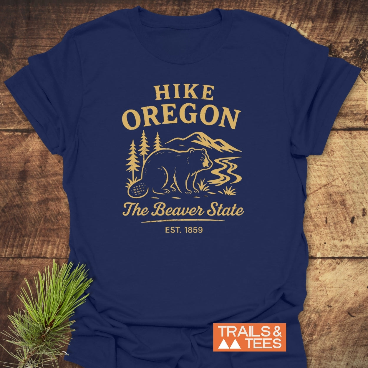 Hike Oregon T-Shirt with a stylized beaver, pine trees, river, and mountains print. Features The Beaver State and EST. 1859. Includes TRAILS & TEES logo and pine sprig for rustic flair.