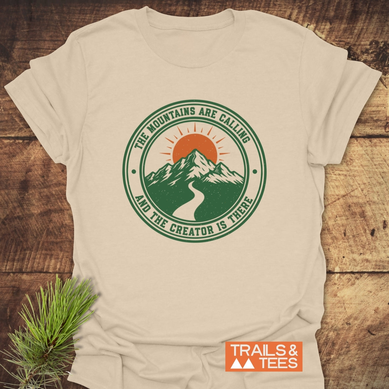 Mountains Are Calling T-Shirt featuring a mountain graphic with sun, surrounded by nature-themed text. Displayed on wood with a pine branch accent, embodying outdoor adventure and comfort.