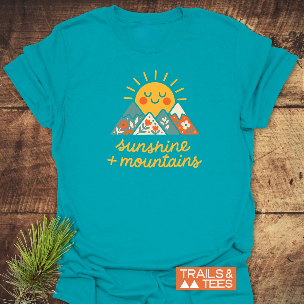 Sunshine And Mountains T-Shirt featuring a cheerful sun and mountain graphic with sunshine + mountains text, on a turquoise shirt. Includes TRAILS & TEES label, emphasizing outdoor adventure and comfort.
