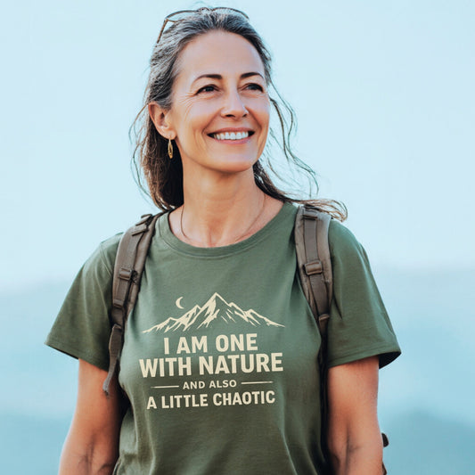 Woman outdoors wearing the One With Nature T-Shirt, showcasing a mountain-and-crescent design with a playful slogan. She carries a backpack, embodying comfort and adventure-ready style.
