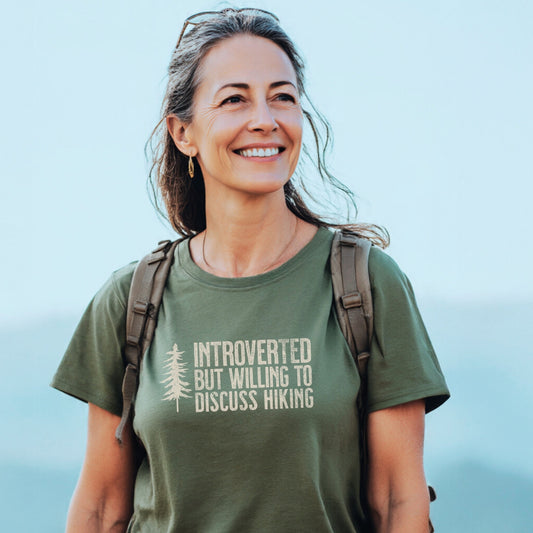 Smiling woman outdoors wearing the Introverted Hiker T-Shirt, featuring the slogan, “INTROVERTED BUT WILLING TO DISCUSS HIKING,” paired with a backpack, embodying trail-ready comfort and adventure spirit.