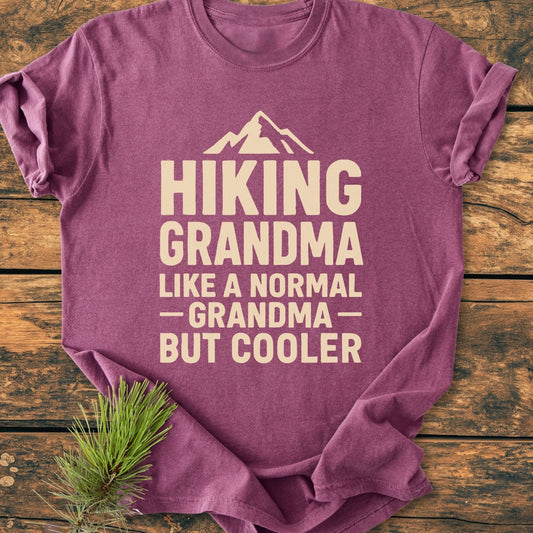 Hiking Grandma Vintage Wash Tee: Purple T-shirt with cream mountain graphic and slogan, 'HIKING GRANDMA, LIKE A NORMAL GRANDMA — BUT COOLER,' styled on wood with pine sprig.