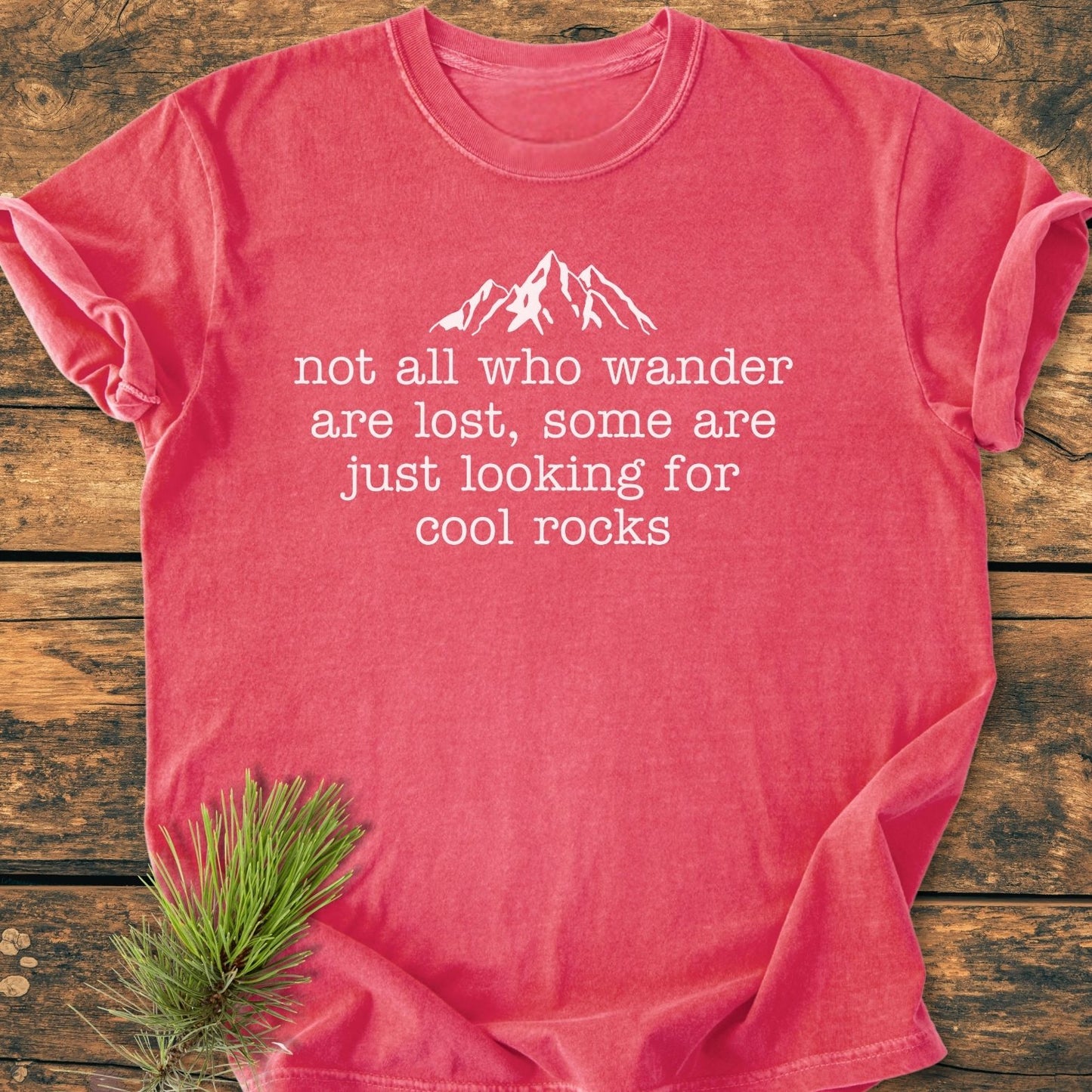 Looking For Cool Rocks Vintage Wash Tee features a coral-red short-sleeve design with a mountain silhouette and quote, laid on wood with pine needles, showcasing its relaxed, durable Comfort Colors fit.