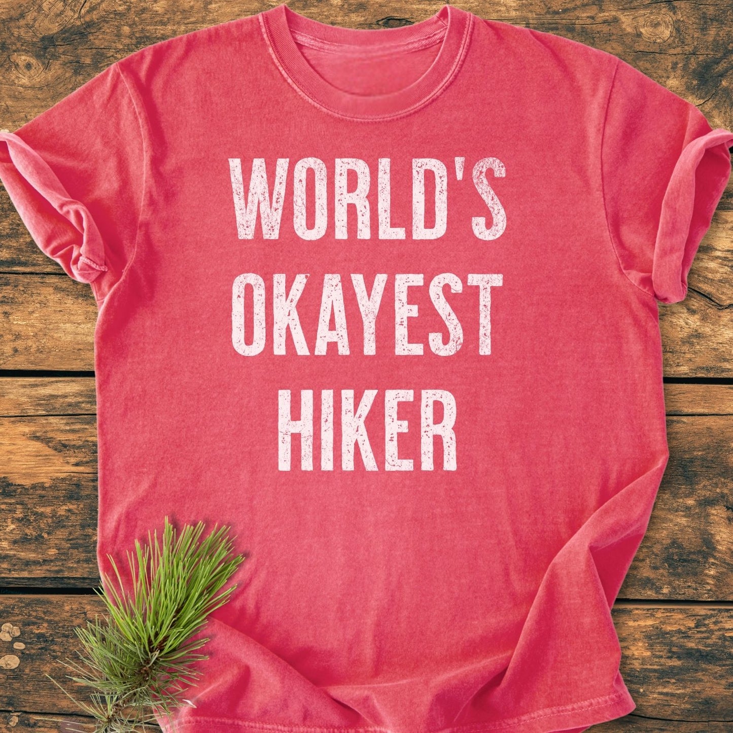 World's Okayest Hiker Vintage Wash Tee, shown flat with a bold white slogan on washed-out red, crafted from soft, garment-dyed cotton. A pine sprig accentuates its rustic, relaxed design.