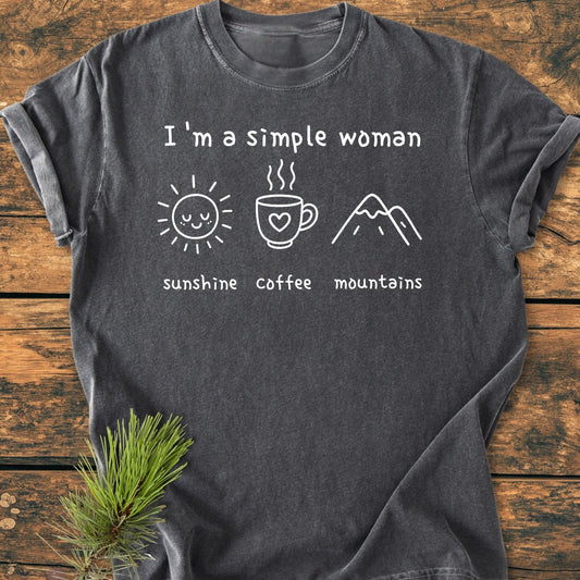 I'm A Simple Woman Vintage Wash Tee on wooden surface, featuring white print of sunshine, coffee, and mountains. Charcoal-gray, 100% ring-spun cotton, relaxed fit, durable stitching, cozy garment-dyed fabric.